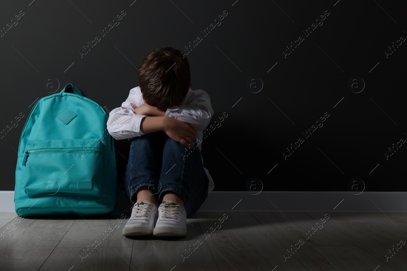 Photo of Upset boy with backpack sitting on floor near black wall, space for text. School bullying