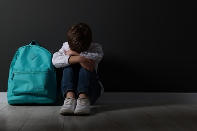 Photo of Upset boy with backpack sitting on floor near black wall, space for text. School bullying