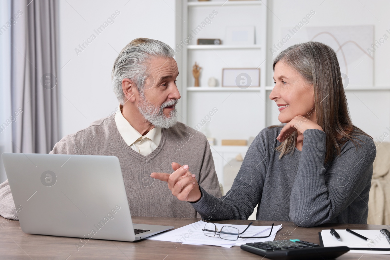 Photo of Elderly couple with papers and laptop discussing pension plan at wooden table in room