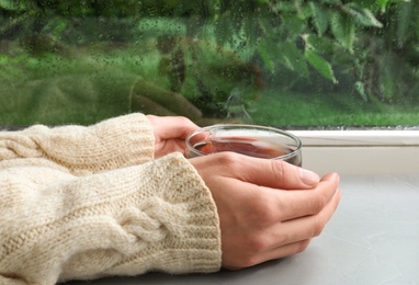 Photo of Woman with cup of hot tea near window on rainy day, closeup