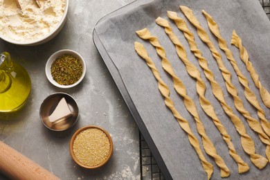 Photo of Baking sheet with homemade breadsticks and ingredients on grey marble table, flat lay. Cooking traditional grissini