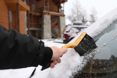 Photo of Man cleaning car windshield from snow with brush outdoors, closeup