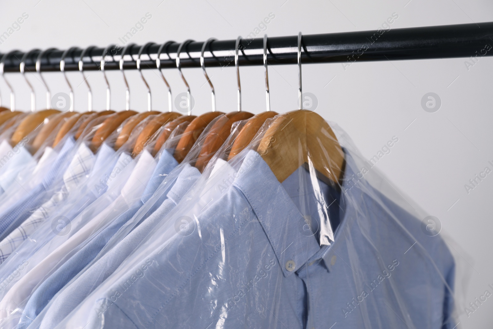 Photo of Hangers with shirts in dry cleaning plastic bags on rack against light background