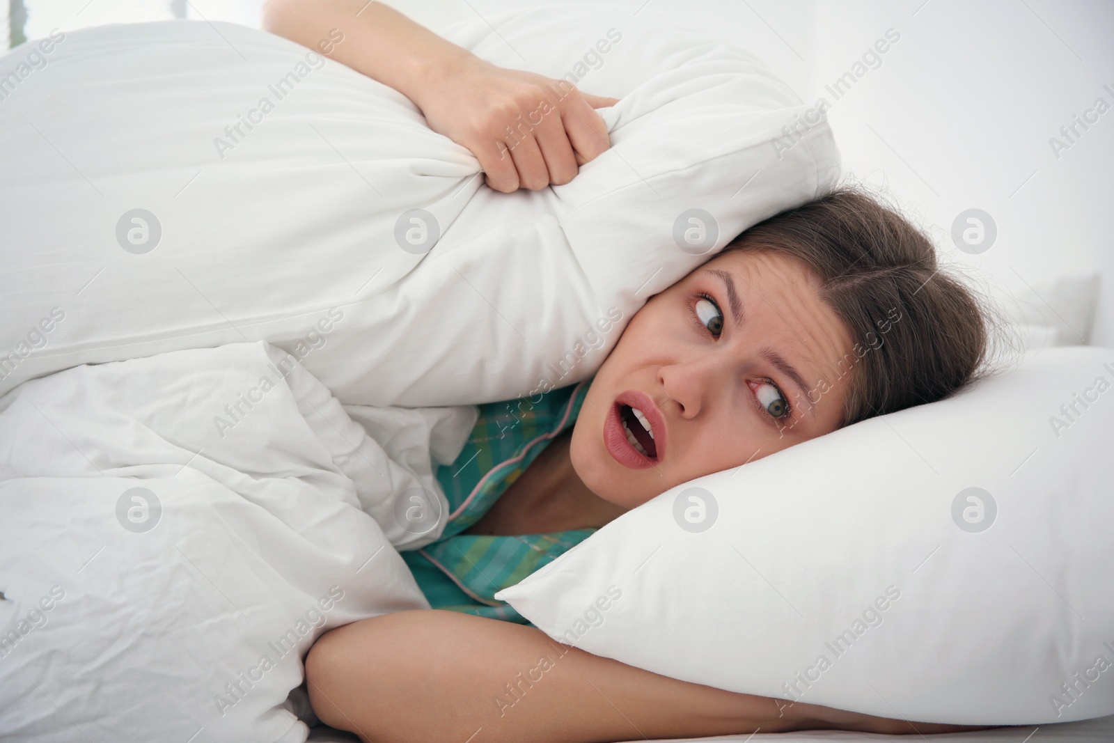 Photo of Emotional young woman covering ears with pillows at home in morning