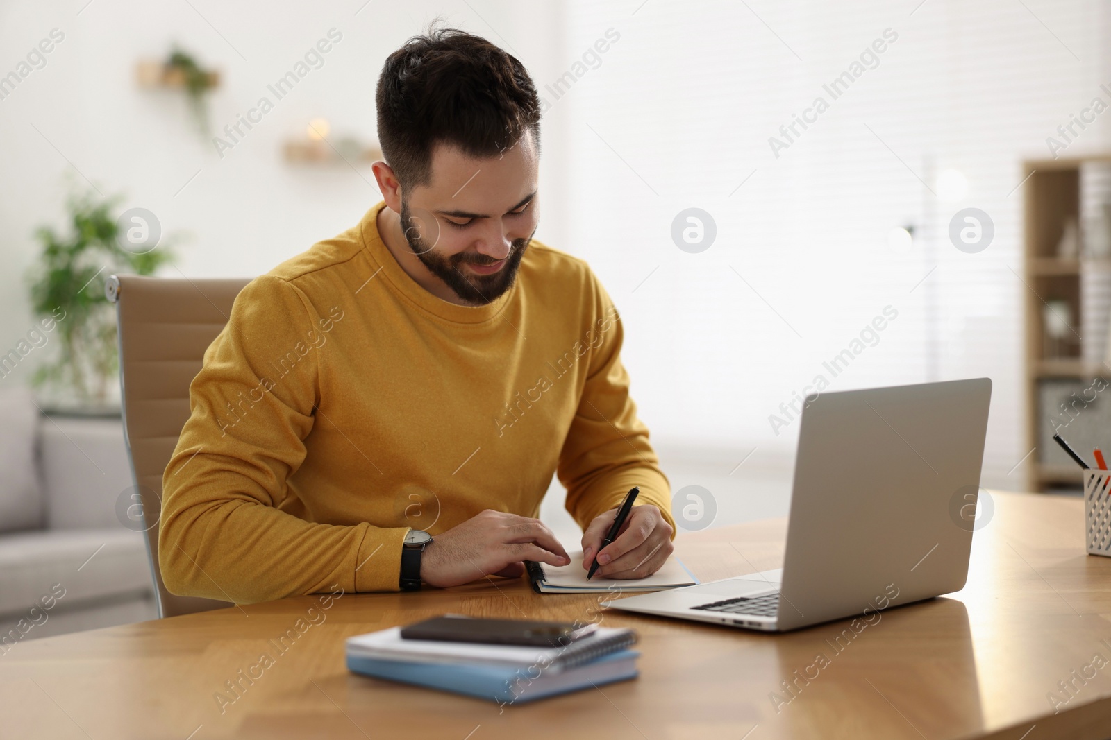 Photo of Young man writing down notes during webinar at table in room