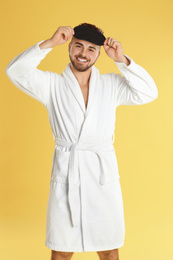 Young man in bathrobe and eye sleeping mask on yellow background