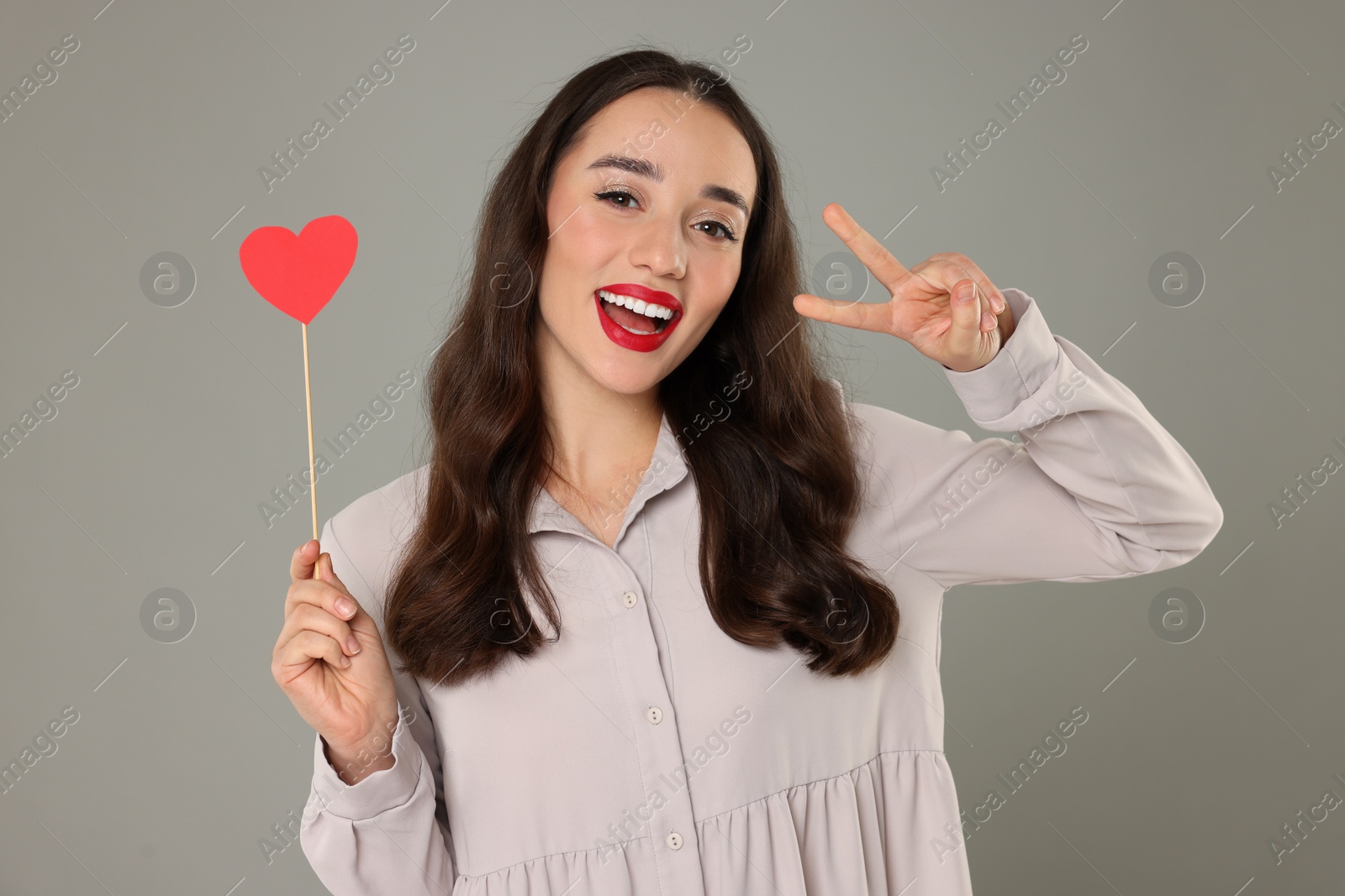 Photo of Beautiful young woman with paper heart showing v-sign on grey background