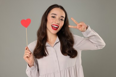 Photo of Beautiful young woman with paper heart showing v-sign on grey background