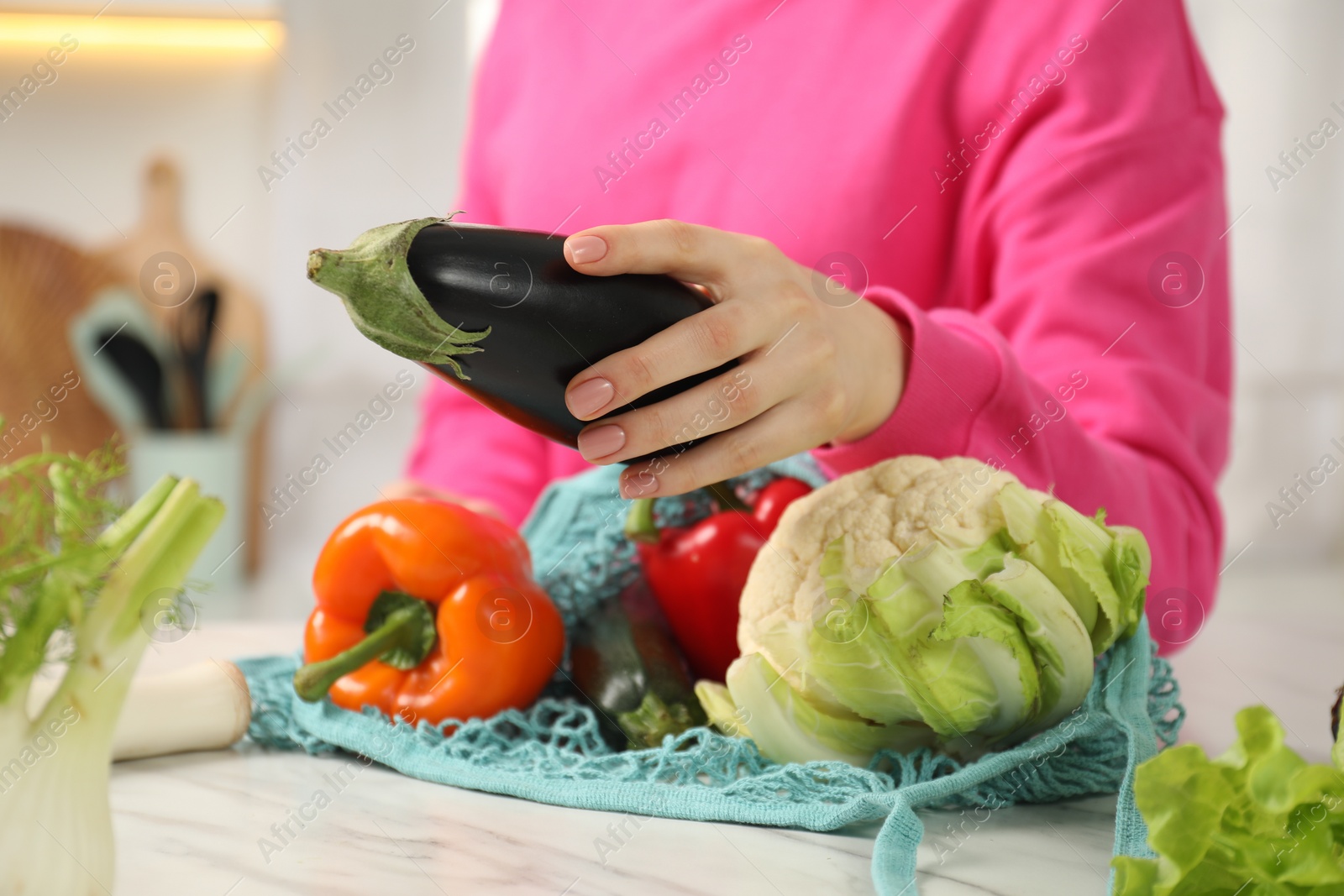 Photo of Woman taking eggplant out from string bag at light marble table, closeup