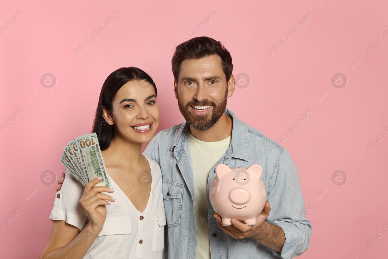 Photo of Happy couple with money and ceramic piggy bank on pale pink background