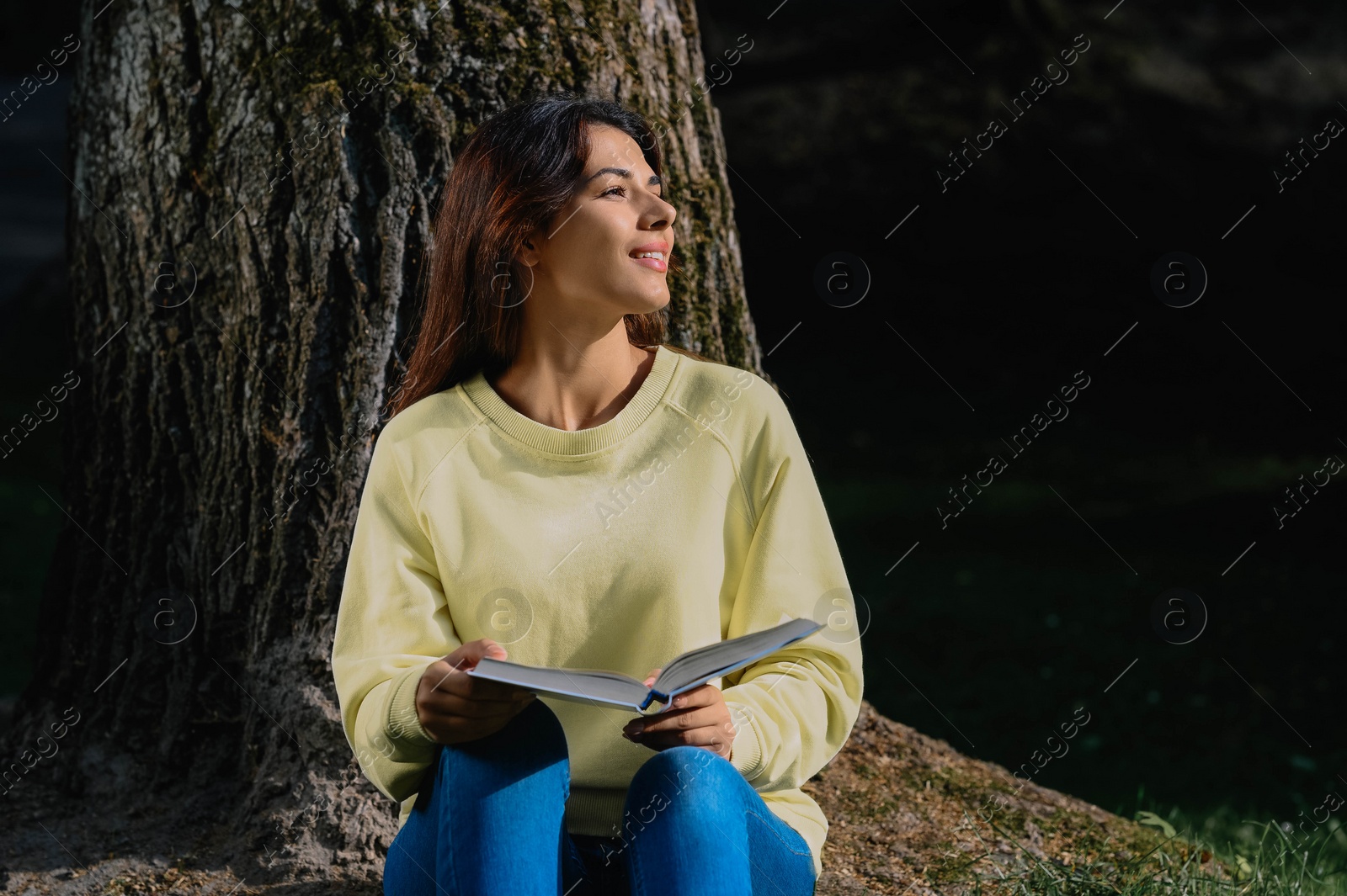 Photo of Young woman reading book near tree in park on sunny day