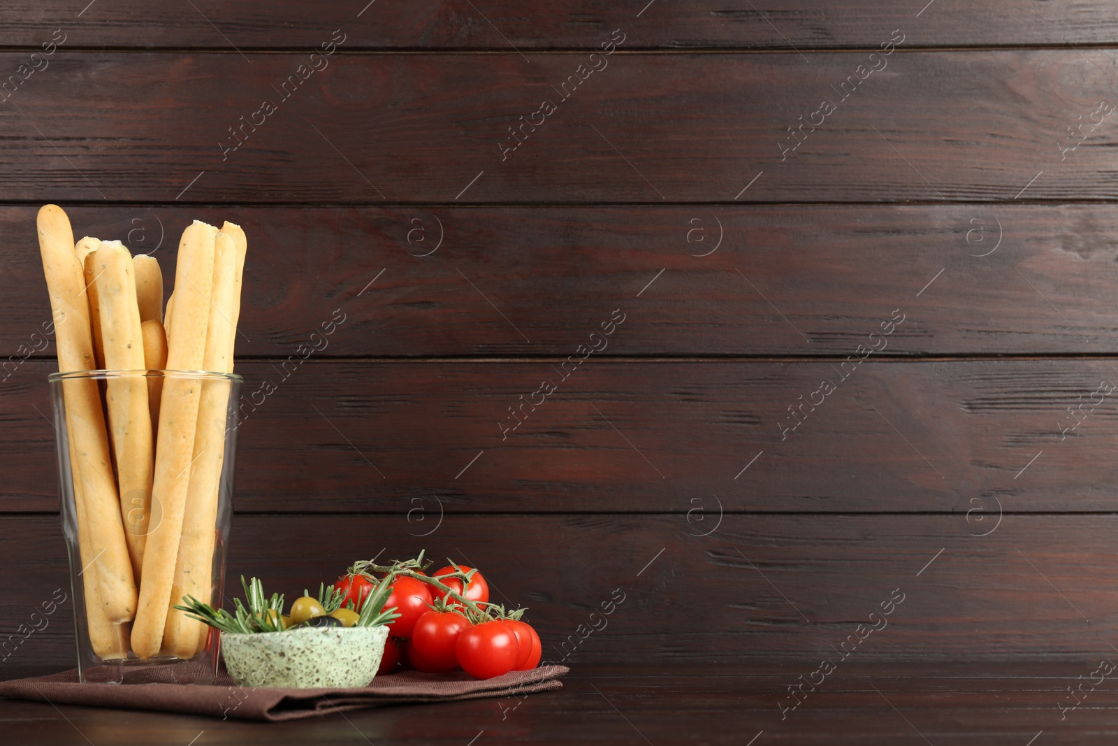 Photo of Fresh delicious grissini in glass, olives with rosemary and tomatoes on wooden table, space for text