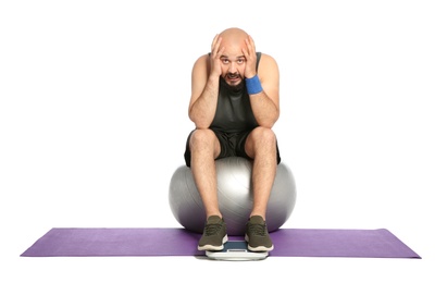 Upset overweight man with scale and fitness ball on white background