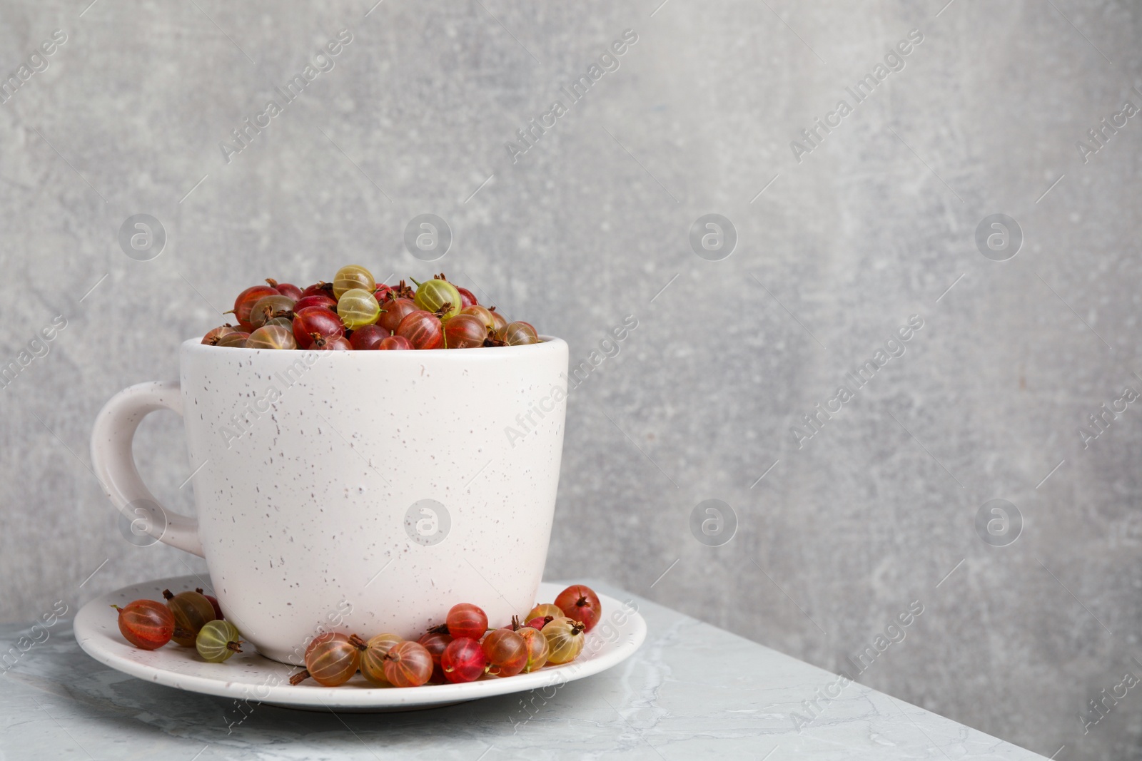 Photo of Cup with ripe gooseberries on light grey marble table, space for text