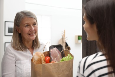 Courier giving paper bag with food products to senior woman indoors
