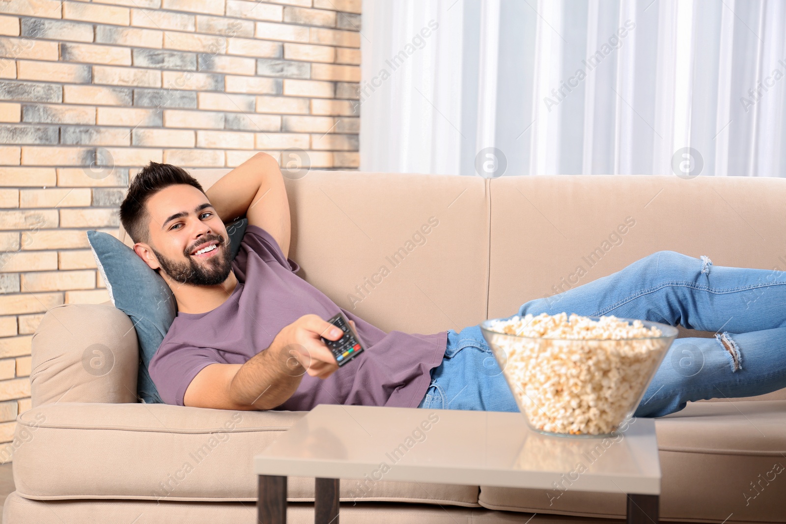 Photo of Young man with remote control and bowl of popcorn watching TV on sofa at home