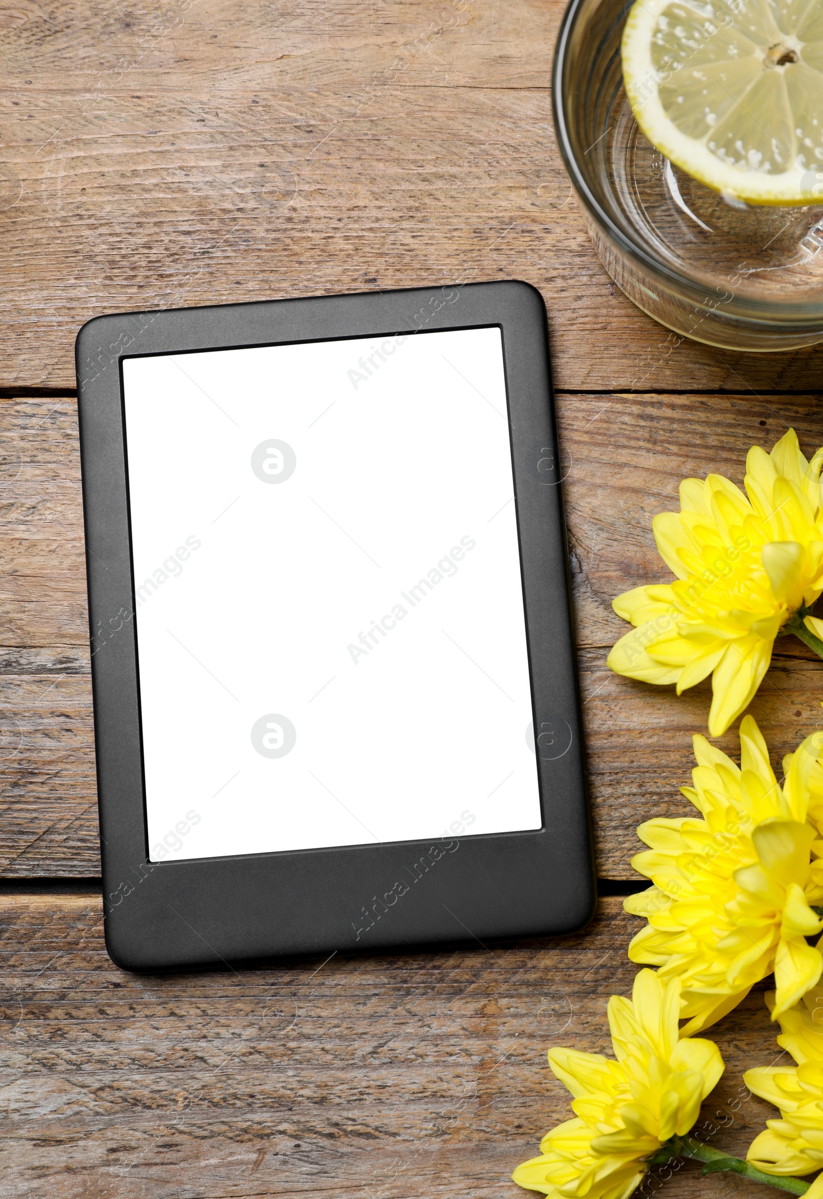 Photo of Modern e-book reader near chrysanthemums and glass of lemon water on wooden table, flat lay