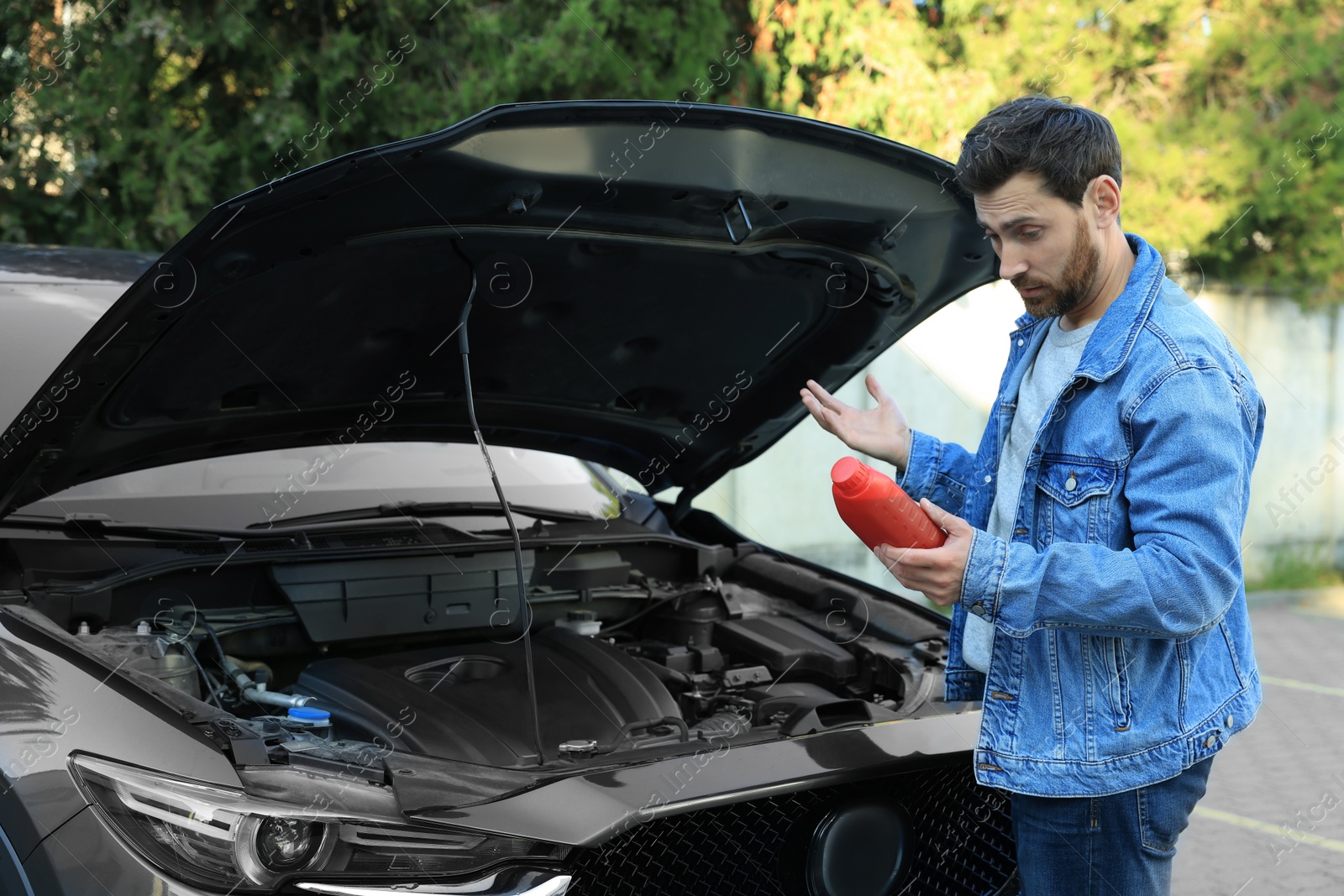 Photo of Puzzled man holding red container of motor oil near car outdoors