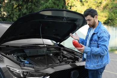 Puzzled man holding red container of motor oil near car outdoors