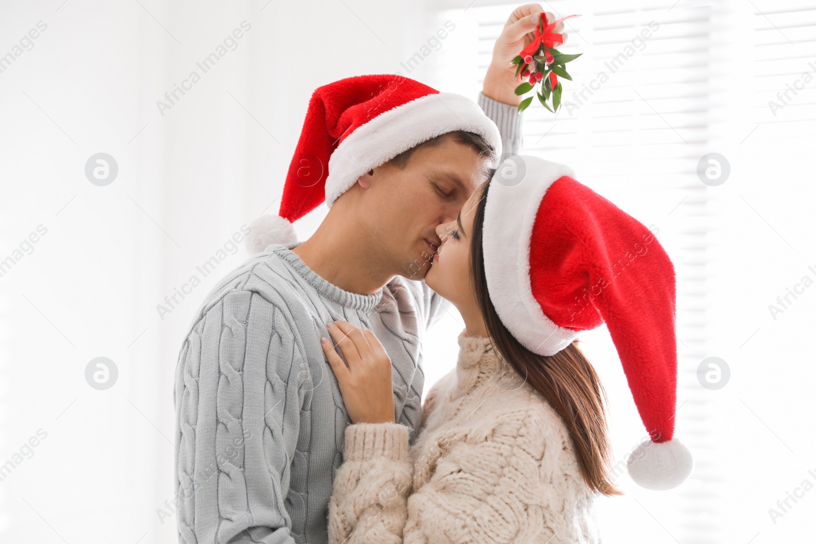 Photo of Happy couple in Santa hats kissing under mistletoe bunch at home