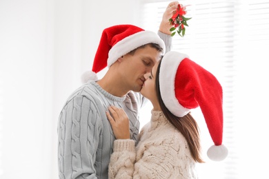 Photo of Happy couple in Santa hats kissing under mistletoe bunch at home