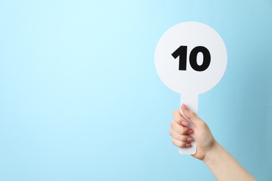 Woman holding auction paddle with number 10 on light blue background, closeup. Space for text