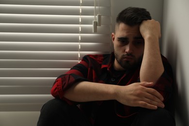 Sad young man near closed blinds at home. Space for text