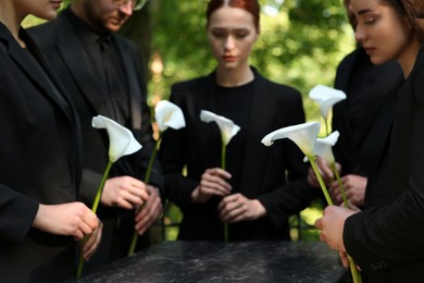 Photo of People with calla lily flowers near granite tombstone at cemetery outdoors, closeup. Funeral ceremony