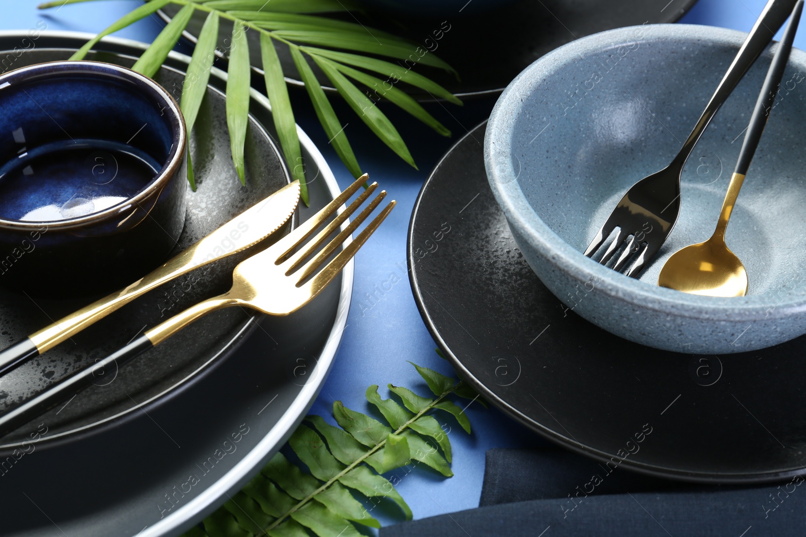 Photo of Stylish ceramic plates, bowls, cutlery and floral decor on blue background, closeup