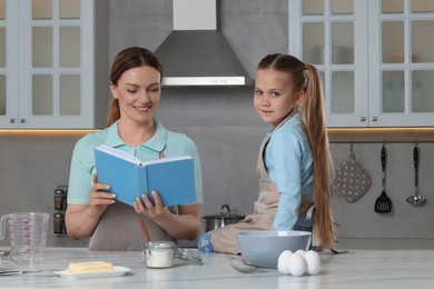 Photo of Cute little girl with her mother cooking by recipe book in kitchen