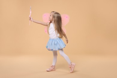 Cute little girl in fairy costume with pink wings and magic wand on beige background