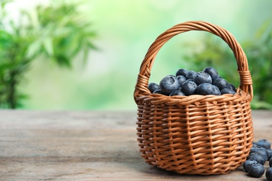 Wicker basket with fresh blueberries on wooden table against blurred green background, space for text