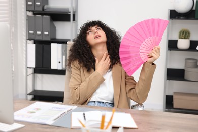 Photo of Young businesswoman waving hand fan to cool herself at table in office
