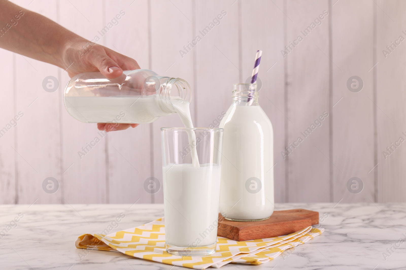 Photo of Woman pouring milk from bottle into glass at white marble table, closeup