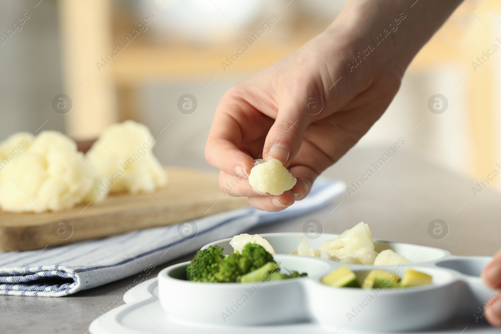Photo of Baby food. Woman putting piece of cauliflower into section plate with different vegetables at grey textured table, closeup