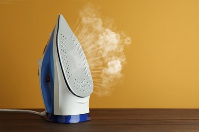 Modern iron with steam on wooden table against orange background, space for text