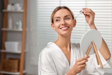 Beautiful woman applying cosmetic serum onto her face in front of mirror in bathroom, space for text