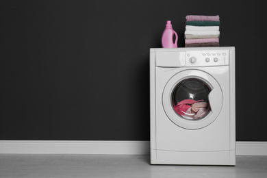 Modern washing machine with laundry, stack of towels and detergent near black wall. Space for text.