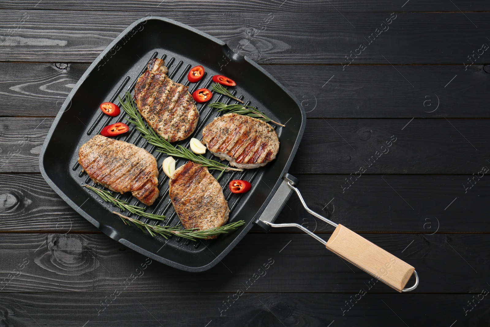 Photo of Grill pan with delicious pork steaks, garlic, chili pepper and rosemary on black wooden table, top view