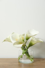 Beautiful calla lily flowers in glass vase on wooden table near white wall