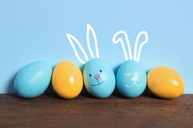 Two eggs with drawn faces and ears as Easter bunnies among others on wooden table against light blue background