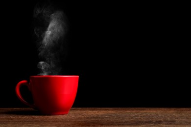 Image of Red cup with hot steaming coffee on wooden table against black background, space for text