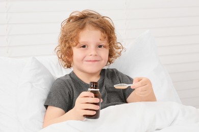 Photo of Cute boy holding measuring spoon with cough syrup in bed. Effective medicine