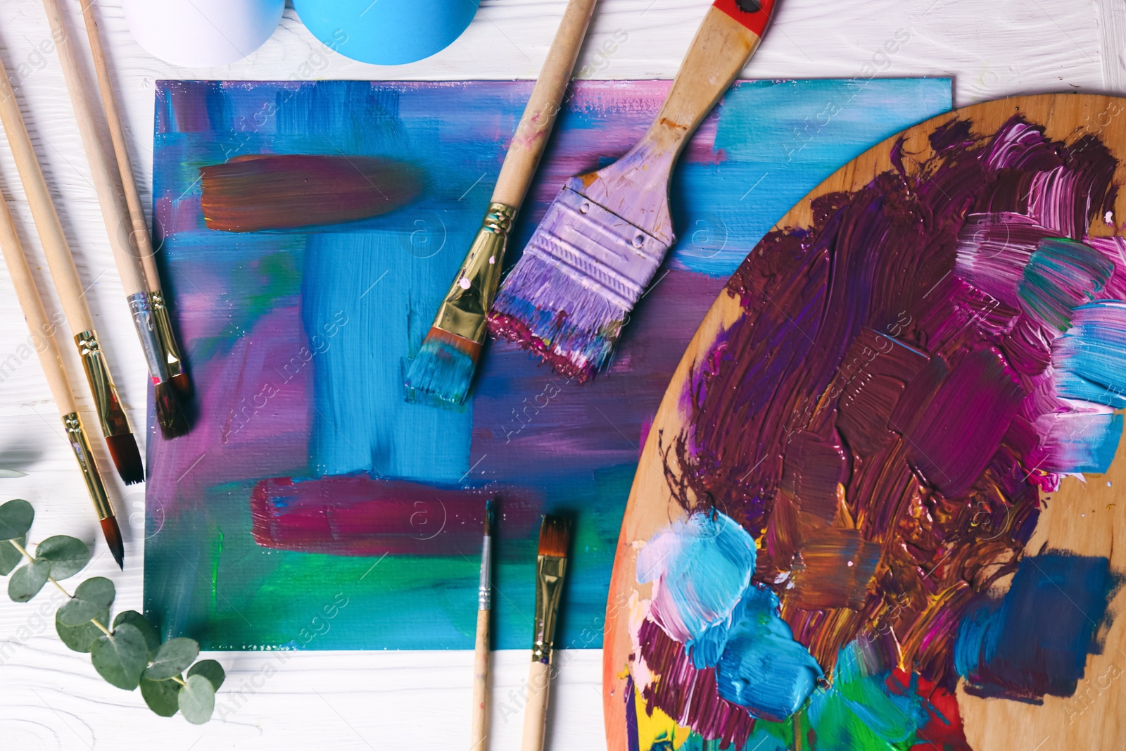 Photo of Canvas with colorful abstract painting, different brushes and wooden artist's palette on white table, flat lay