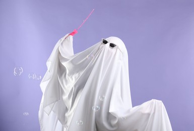 Photo of Funny ghost. Person in white sheet, sunglasses and soap bubbles on violet background