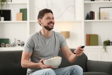 Happy man with popcorn changing TV channels with remote control on sofa at home