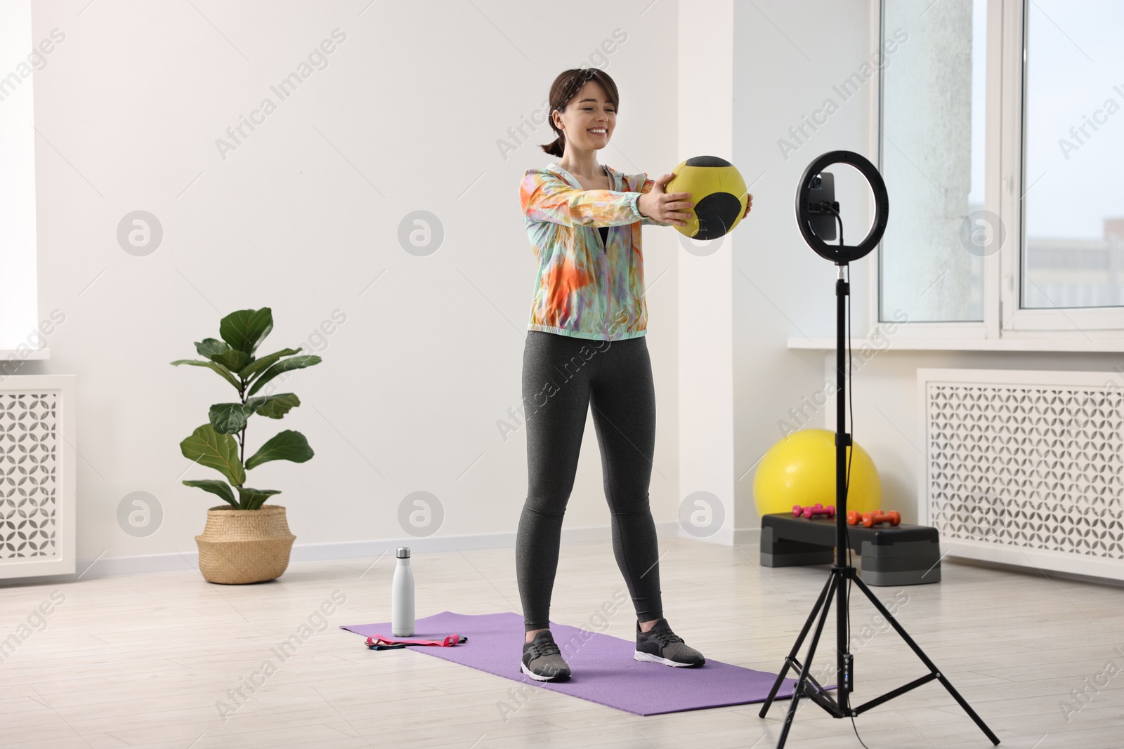 Photo of Happy sports blogger training with medicine ball while streaming online fitness lesson with smartphone at home