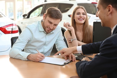 Photo of Young couple and salesman sitting at table in car salon. Buying auto