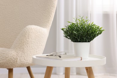 Photo of Potted artificial plant, book and glasses on side table near armchair indoors