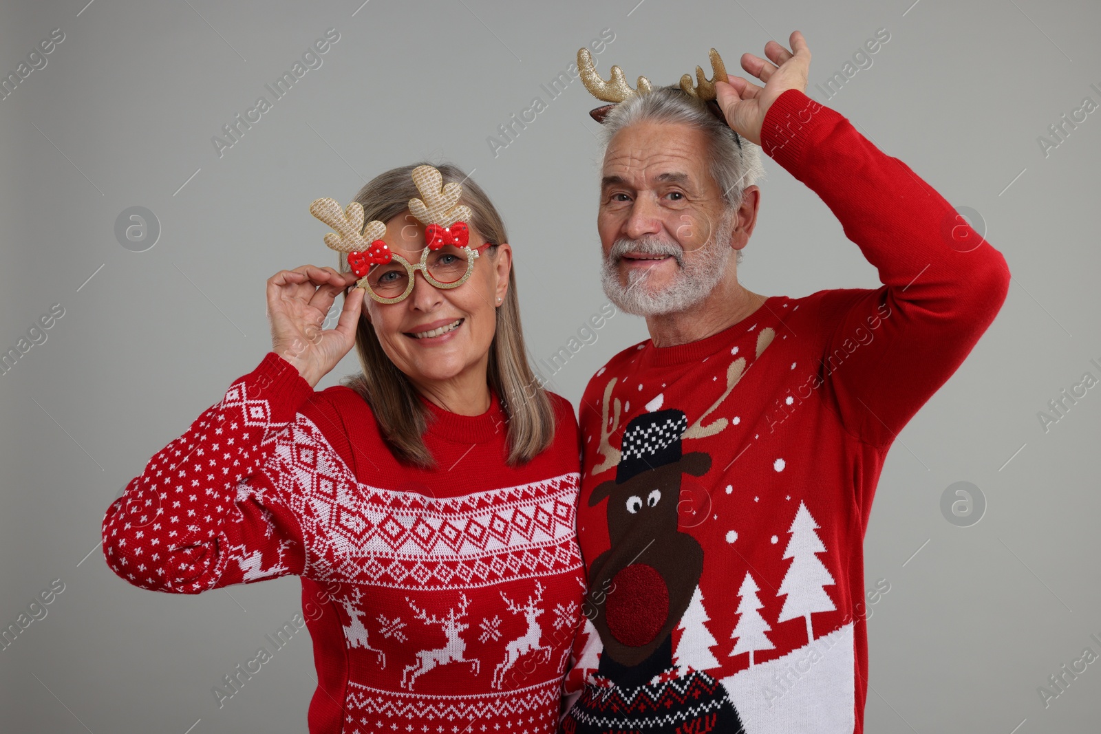 Photo of Senior couple in Christmas sweaters, reindeer headband and funny glasses on grey background